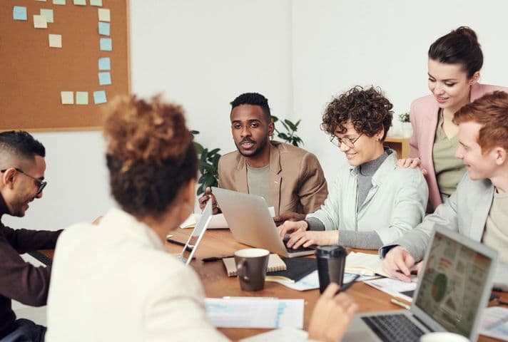 A diverse group of six people are gathered around a table in a meeting room. They are engaged in a discussion about SEO with laptops, papers, and coffee cups in front of them.