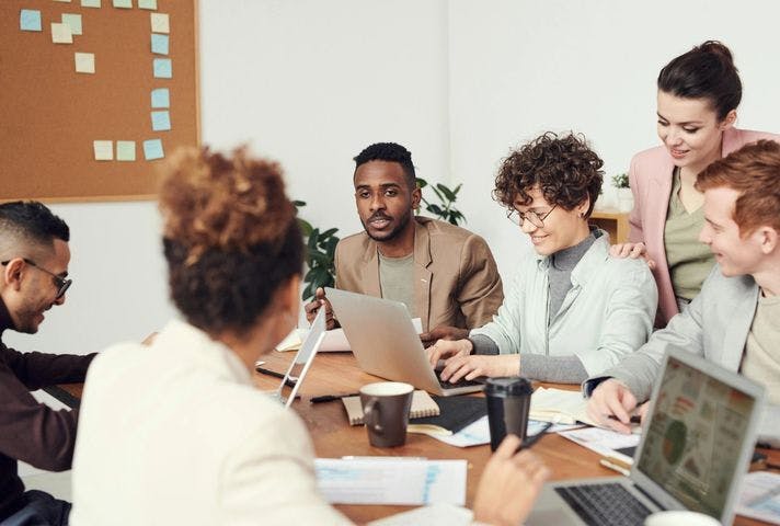 A diverse group of six people are gathered around a table in a meeting room. They are engaged in a discussion about SEO with laptops, papers, and coffee cups in front of them.