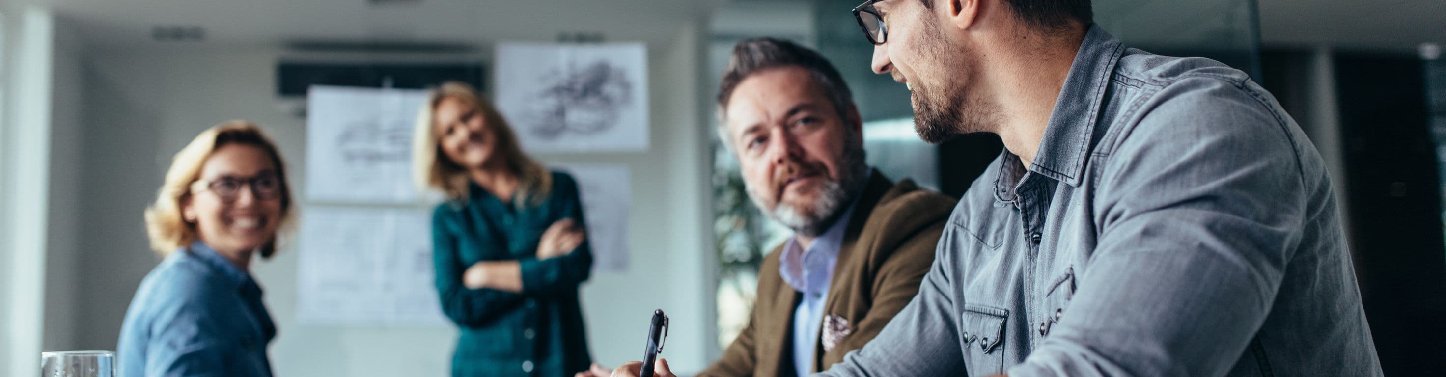 Stock photo of four people are sitting around a table in a office engaged in a meeting. Papers and a glass of water lay on the table.