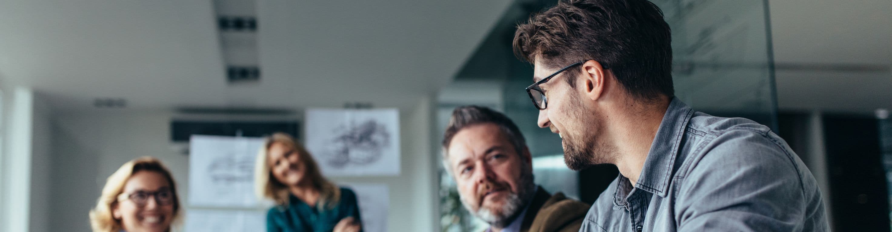 Stock photo of four people are sitting around a table in a office engaged in a meeting. Papers and a glass of water lay on the table.