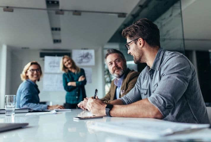 Stock photo of four people are sitting around a table in a office engaged in a meeting. Papers and a glass of water lay on the table.