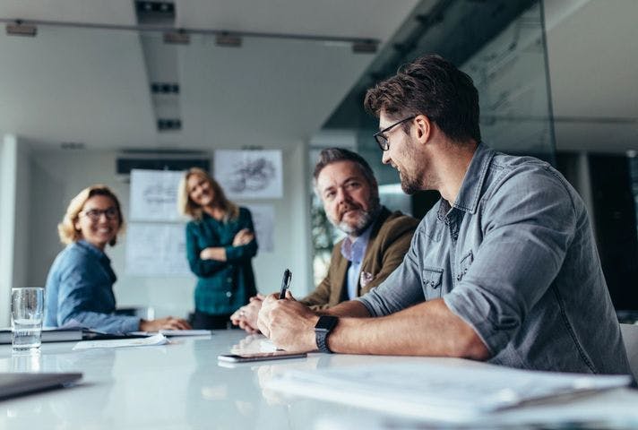 Stock photo of four people are sitting around a table in a office engaged in a meeting. Papers and a glass of water lay on the table.