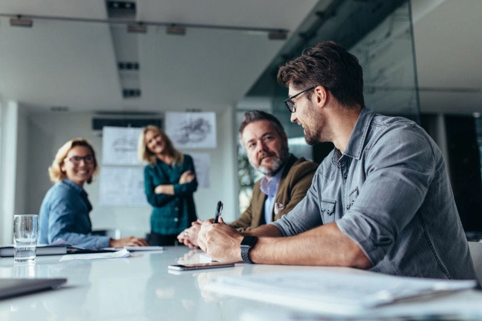 Stock photo of four people are sitting around a table in a office engaged in a meeting. Papers and a glass of water lay on the table.