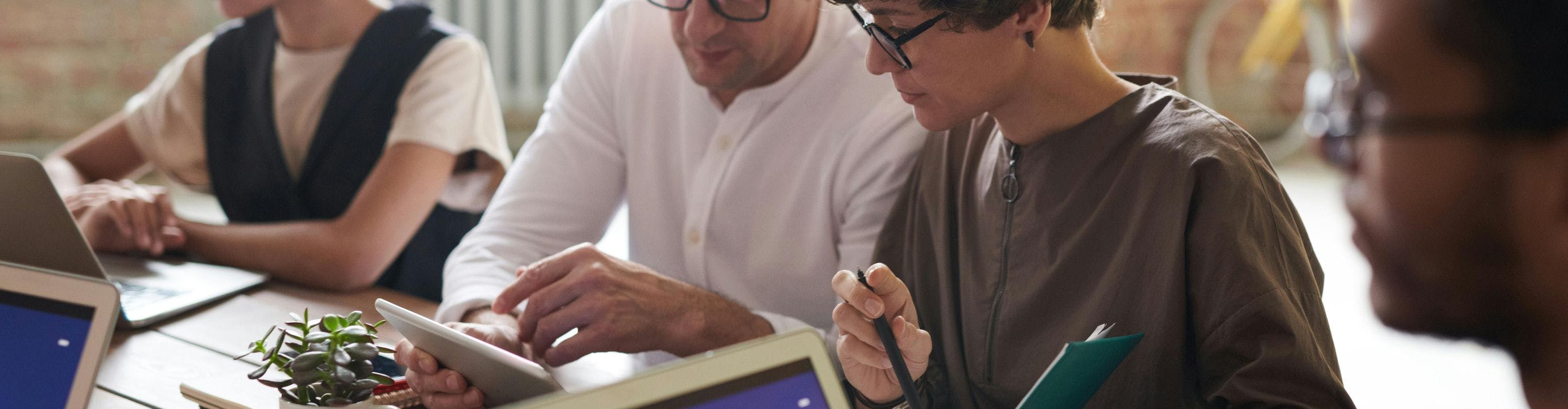 A group of people sits around a table working on laptops. Two individuals in the foreground closely examine a tablet, while another person takes notes.