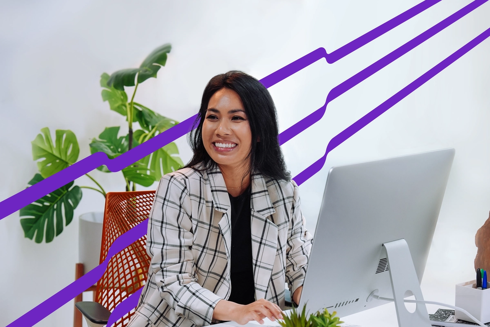 A woman sits at a desk, smiling while looking at a computer monitor. The desk features notepads, a pen, and a lush plant. Purple diagonal lines extend in the background.