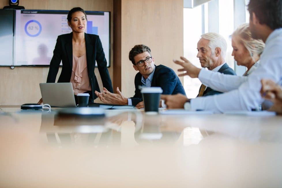 A group of professionals sit around a conference table in an office engaged in a discussion. A woman is presenting and stands by a screen.