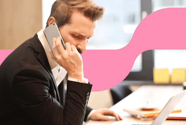 Stock photo of a man in a black suit seated at a desk, holding a phone to his ear with a laptop and a cup of coffee in front of him. A pink wave design overlays part of the background.