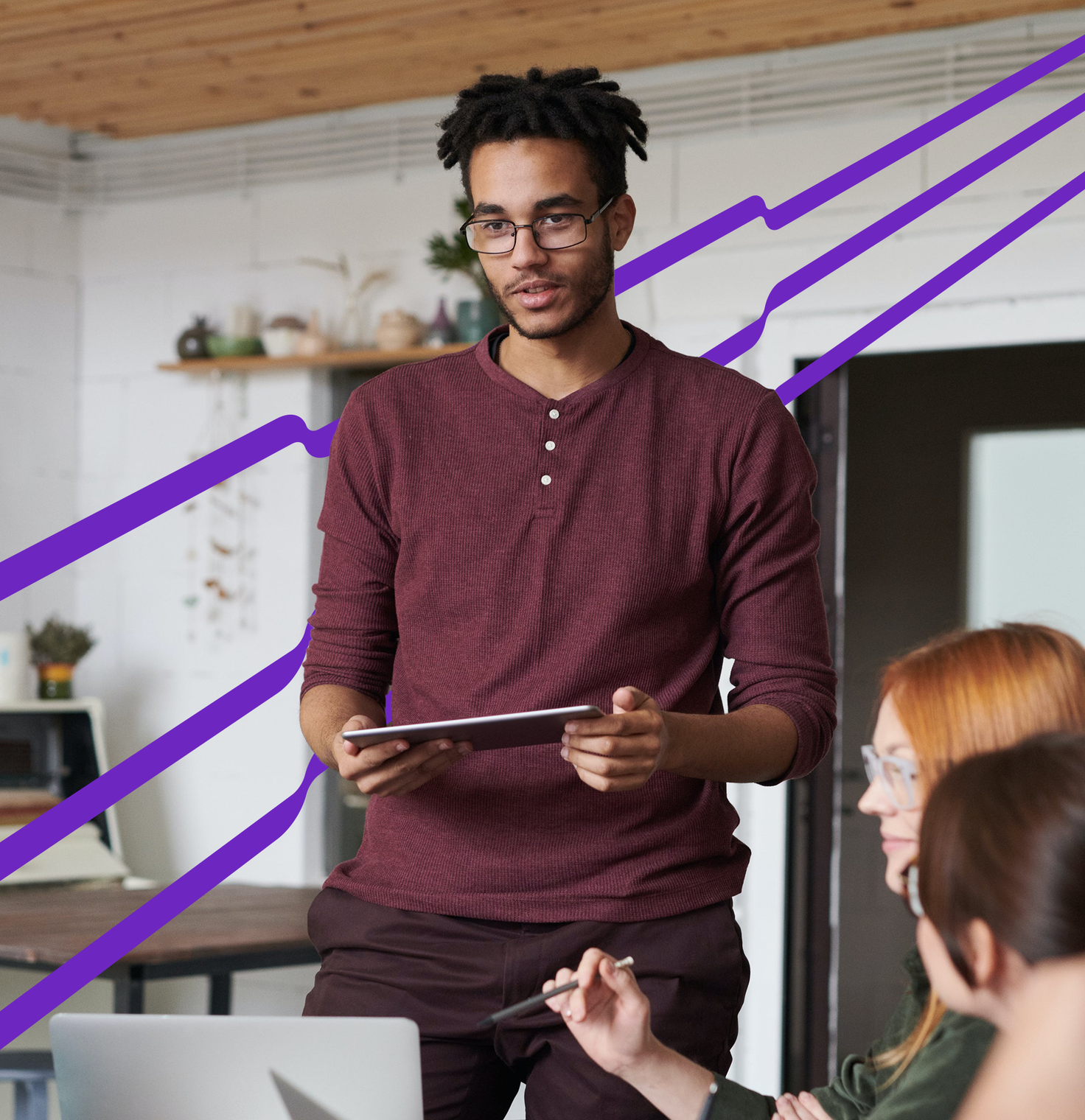 Stock photo of a man stands holding a tablet and speaks to a group of people in a modern office setting with purple lines extending across the picture.