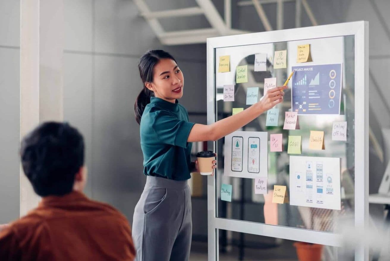 Woman on a presentation board explaining graphs and charts to a man sitting nearby