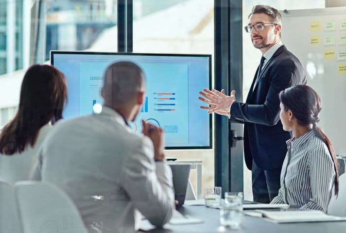 A man in a suit stands in front of a screen displaying charts while addressing three colleagues in an office.