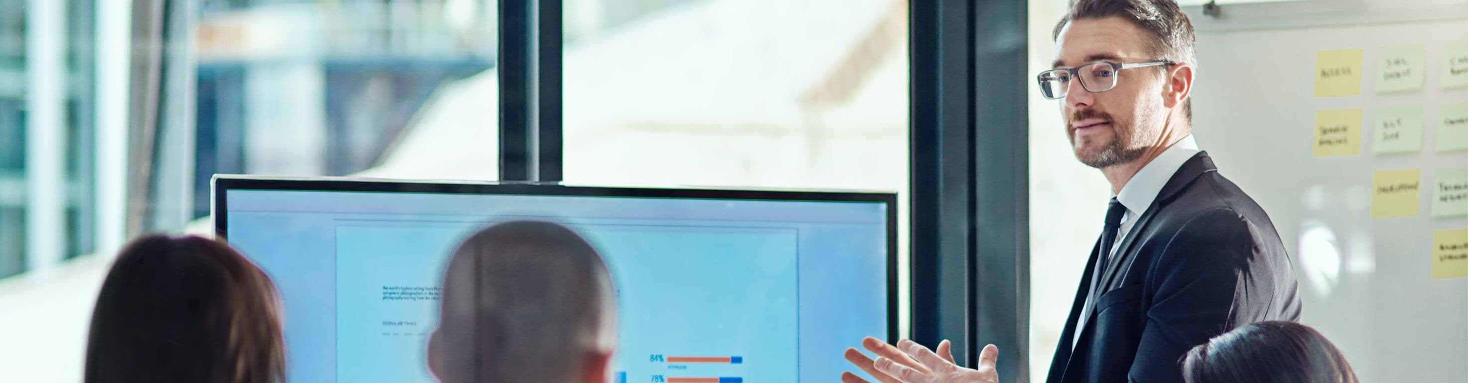 A man in a suit stands in front of a screen displaying charts while addressing three colleagues in an office.