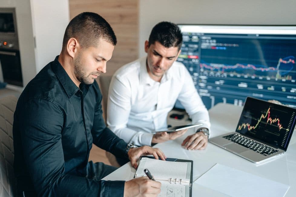 Two men are seated at a desk in an office. One in a black shirt is writing in a notebook, while the other in a white shirt holds a tablet. A laptop and a large monitor display financial data.