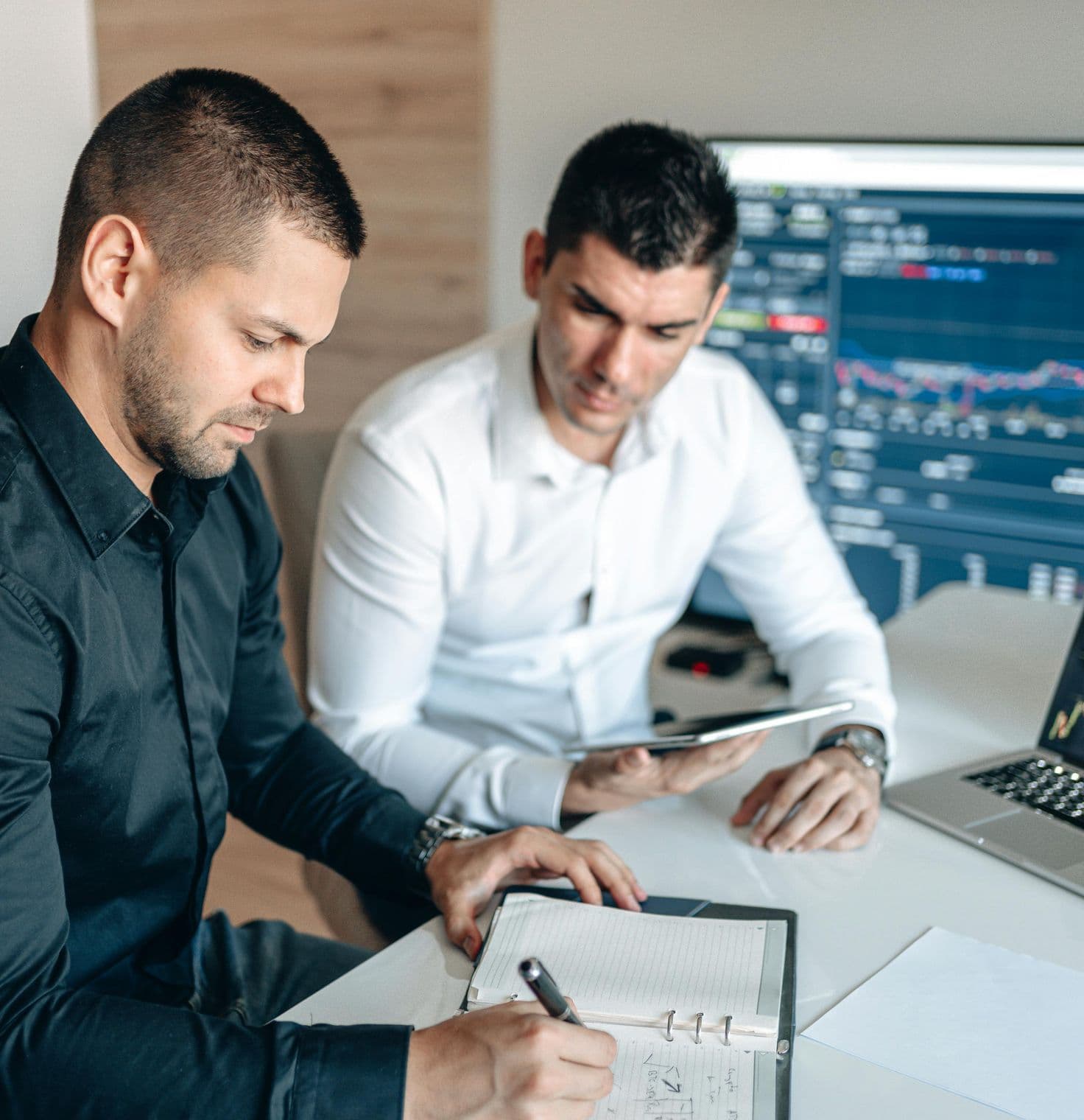 Two men are seated at a desk in an office. One in a black shirt is writing in a notebook, while the other in a white shirt holds a tablet. A laptop and a large monitor display financial data.