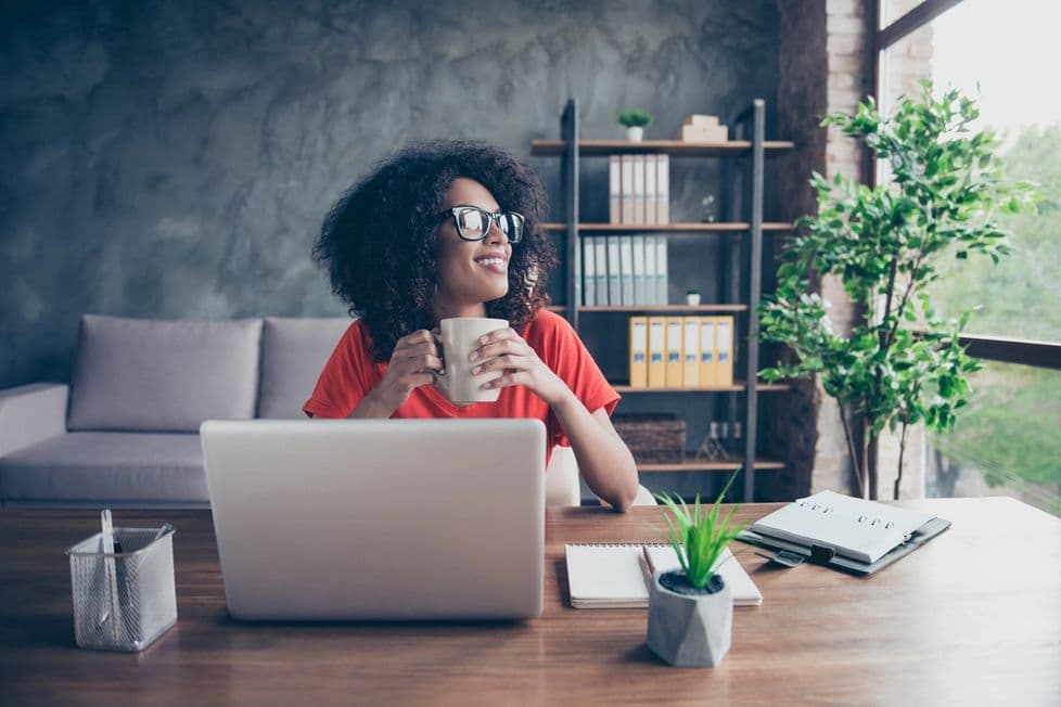 Stock photo of a woman sitting at a wooden desk with a laptop, notepad, plant, and other office supplies in the room. A bookshelf is featured in the background.