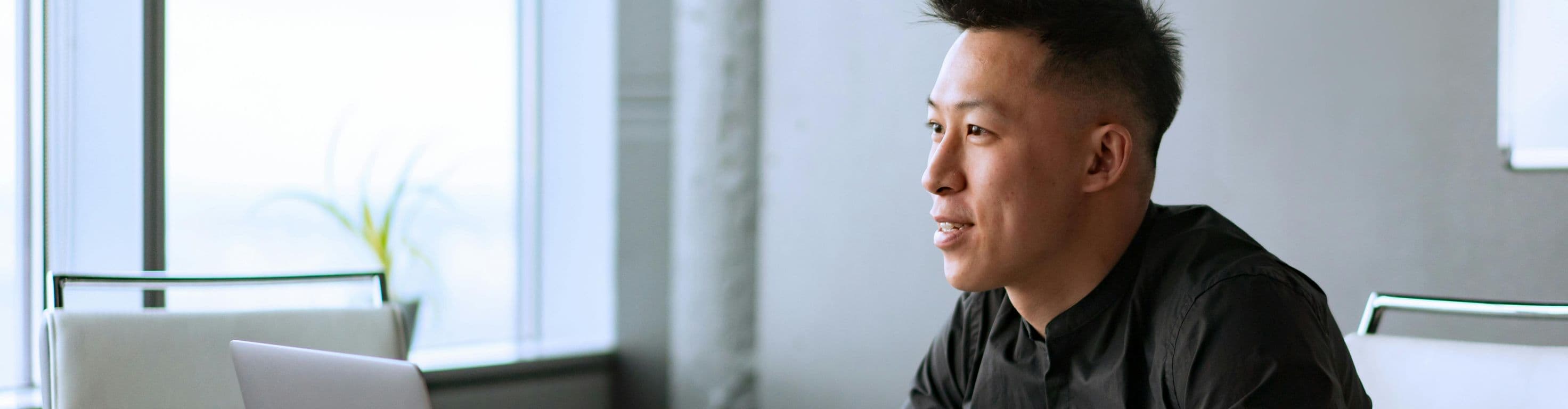 A man in a black shirt is seated at a desk in a home office with a laptop in front of him. He's writing in a notebook as he reviews the content on his screen.