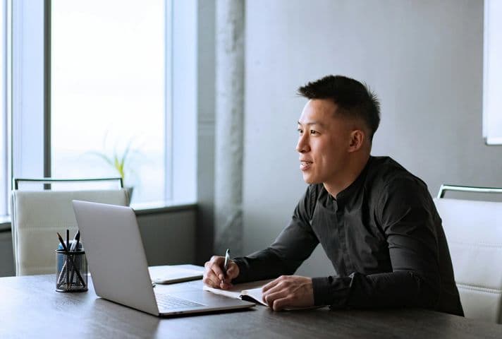 A man in a black shirt is seated at a desk in a home office with a laptop in front of him. He's writing in a notebook as he reviews the content on his screen.