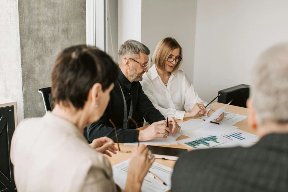 A group of four people sit around a table in an office room. They are reviewing various documents and charts showing some analytics.