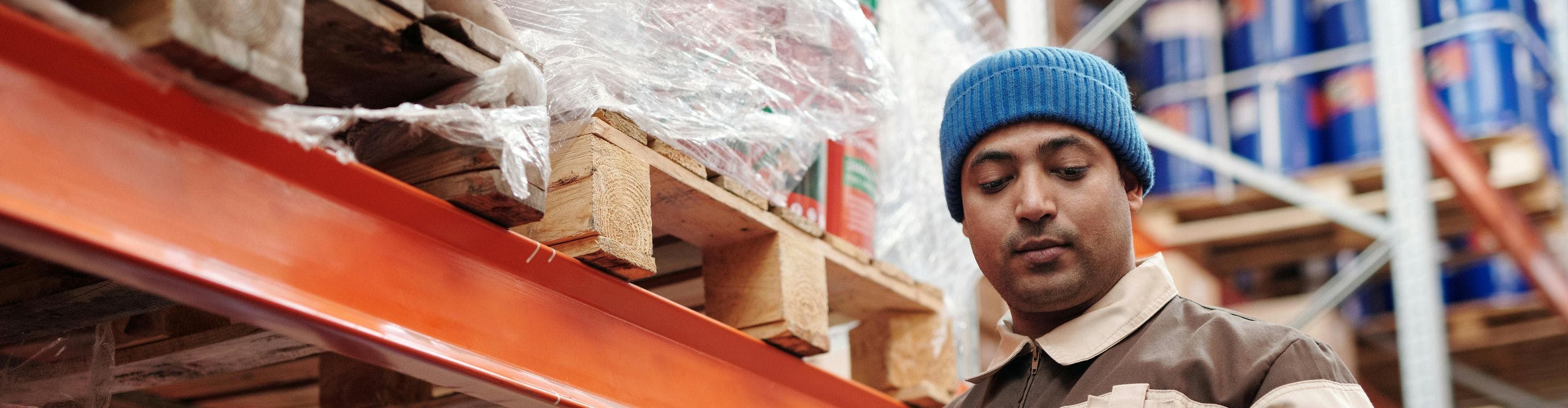 A worker wearing a blue beanie and a brown and beige uniform is holding a tablet and standing beside shelves in a warehouse. Several large containers and pallets are visible around him.