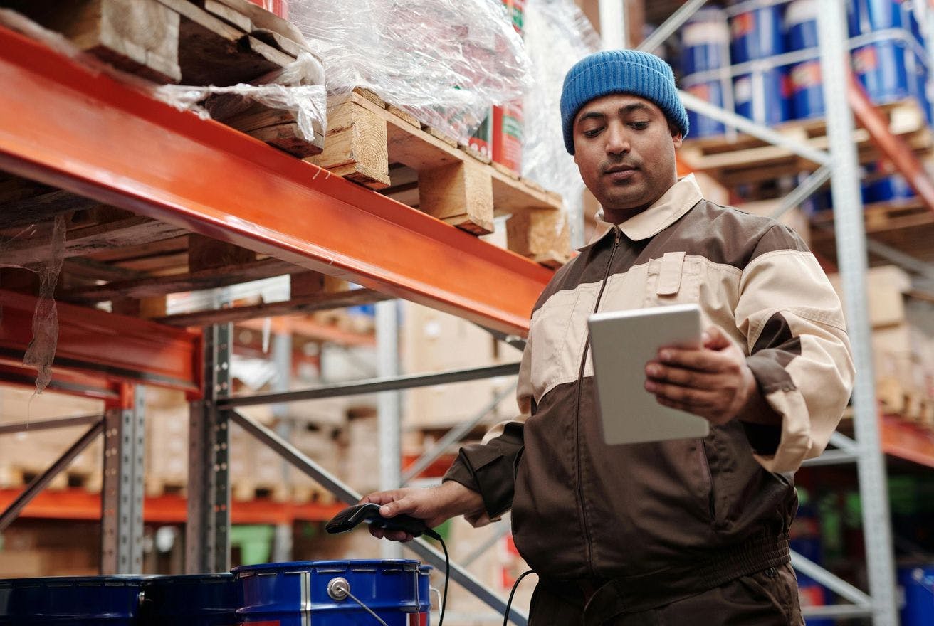 A worker wearing a blue beanie and a brown and beige uniform is holding a tablet and standing beside shelves in a warehouse. Several large containers and pallets are visible around him.