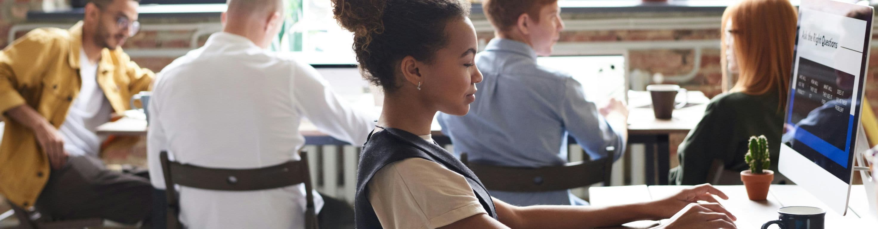 A woman and a group of people are working at desks in a open office space. The workspace has a few desks and monitors.