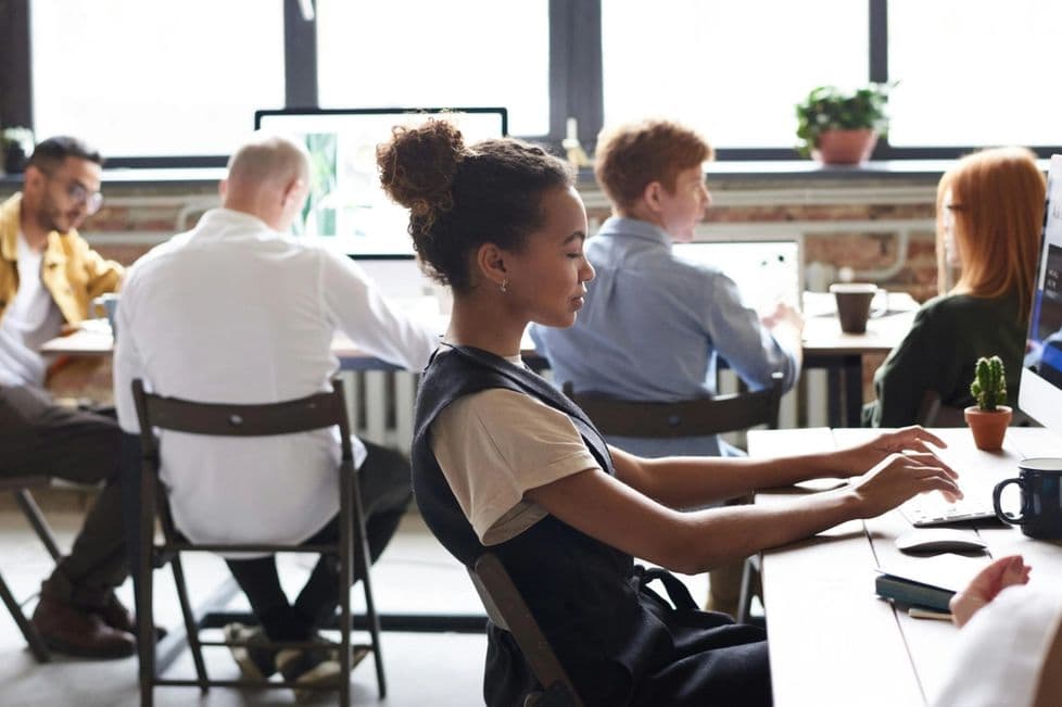 A woman and a group of people are working at desks in a open office space. The workspace has a few desks and monitors.