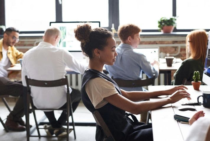 A woman and a group of people are working at desks in a open office space. The workspace has a few desks and monitors.