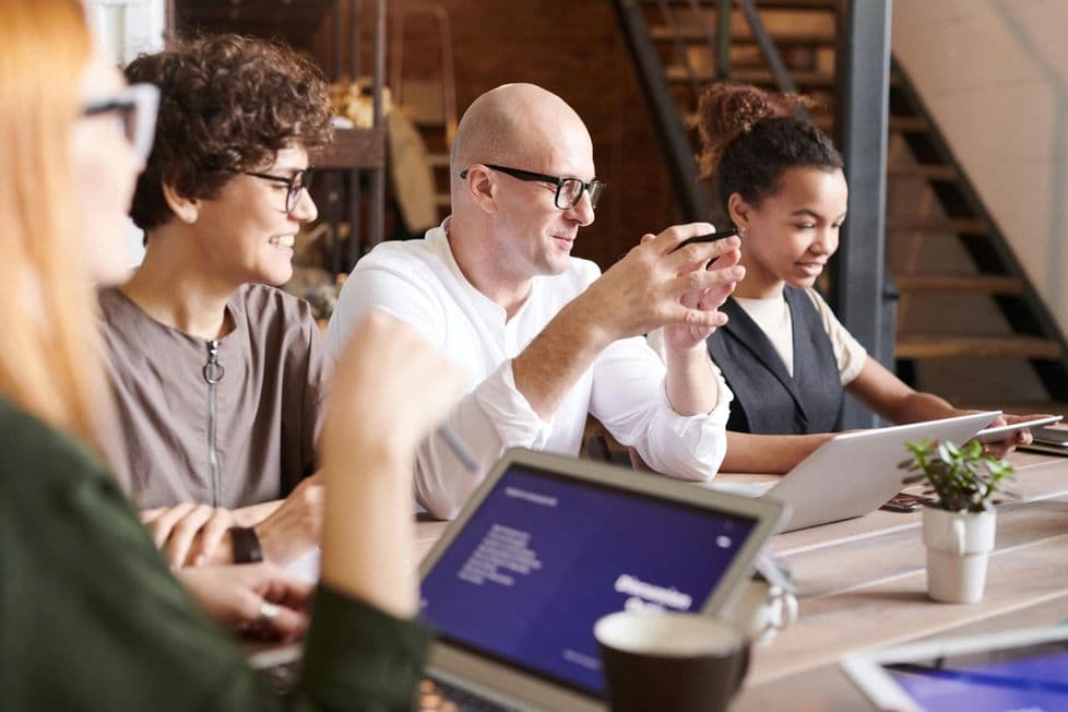 A diverse group of four people sits around a table, engaged in a discussion while working on laptops.